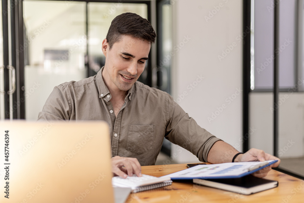 A young businessman sitting in the office with a laptop, is pressing a calculator to calculate the balance, he founded a startup, he is a young entrepreneur who has the skills to run a growing company A young businessman sitting in the office with a laptop, is pressing a calculator to calculate the balance, he founded a startup, he is a young entrepreneur who has the skills to run a growing company