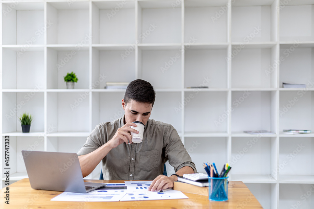 Young businessman sitting in the office with a laptop, taking a hot coffee break while checking documents, he founded a startup, he is a young entrepreneur who has the skills to run a growing company.