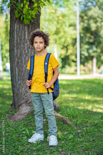 Little schoolboy with curly hair in a yellow t-shirt stands alone in the park