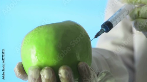 Close-up of a human in a medical coat and gloves injecting a syringe into an apple with some liquid on a blue background