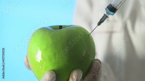 Close-up of a human in a medical coat and gloves injecting a syringe into an apple with some liquid on a blue background