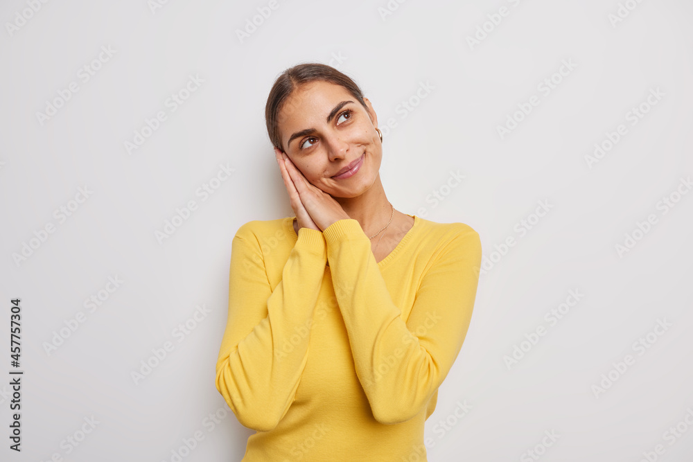 Dreamy brunette woman leans on palms has gentle expression thinks about something positive wears casual yellow jumper isolated over white background. Romantic female model ponders on future. Dreamy brunette woman leans on palms has gentle expression thinks about something positive wears casual yellow jumper isolated over white background. Romantic female model ponders on future.