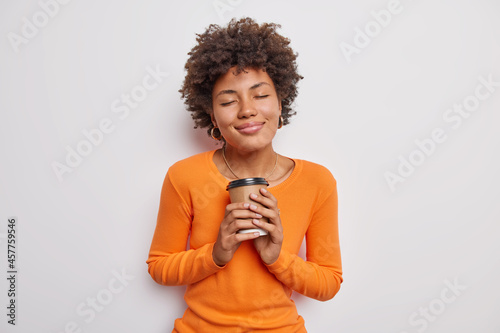 Pleased African American woman with curly hair holds paper disposable cup of coffee closes eyes from satisfaction wears casual orange jumper isolated over white background. People and drinking