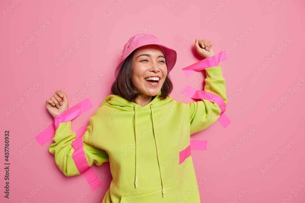 Positive Asian woman laughs happily plastered to wall with sticky tapes wears panama and hoodie feels not free isolated over pink background. Studio shot. Optimistic eastern female in captivity Positive Asian woman laughs happily plastered to wall with sticky tapes wears panama and hoodie feels not free isolated over pink background. Studio shot. Optimistic eastern female in captivity