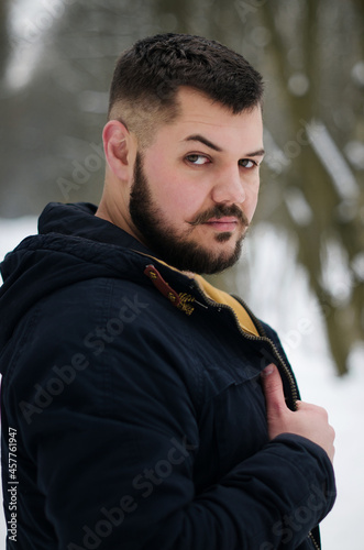 A young man with mustaches and raised eyebrow holds his hand over his jacket and walks in a pine forest. Portrait of a brutal bearded man. Winter forest tourism concept