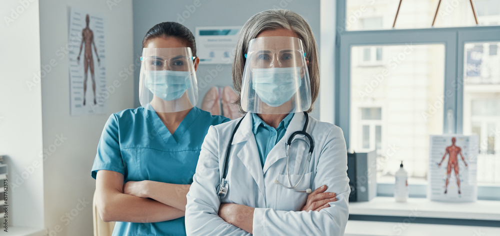 Two confident women coworkers in medical uniform and protective workwear keeping arms crossed and looking at camera while working in the hospital - obrazy, fototapety, plakaty 