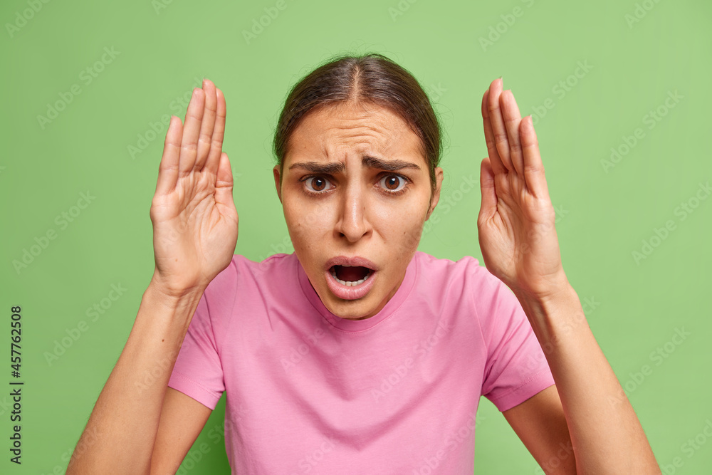 Indoor shot of frustrated young woman raises palms looks unhappily has discontent worried expression frowns face wears casual pink t shirt isolated over green background. Negative emotions concept Indoor shot of frustrated young woman raises palms looks unhappily has discontent worried expression frowns face wears casual pink t shirt isolated over green background. Negative emotions concept