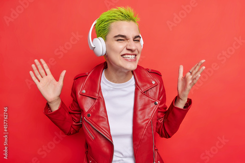 Foto Waist up shot of happy hipster girl clenches teeth smiles gladfully at camera has fun while listening music in headphones wears leather jacket isolated over bright red background