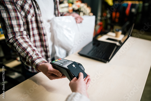 Close-up shopper hand gives credit card to pay through payment terminal, seller behind counter makes payment and gives goods in box to buyer. Pay use non-cash technology © Elizaveta