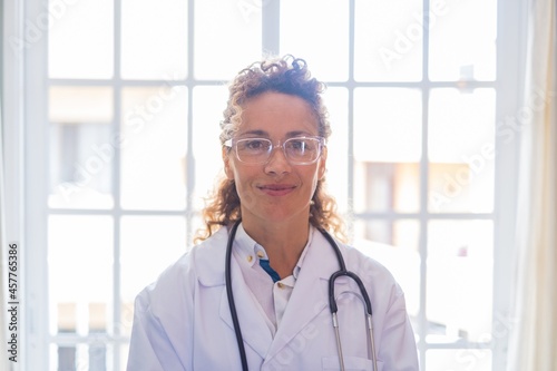 Portriat of young female doctor in uniform and stethoscope standing against window at clinic. Confident frontline medical practicitioner or healthcare worker in eyeglasses at hospital. photo