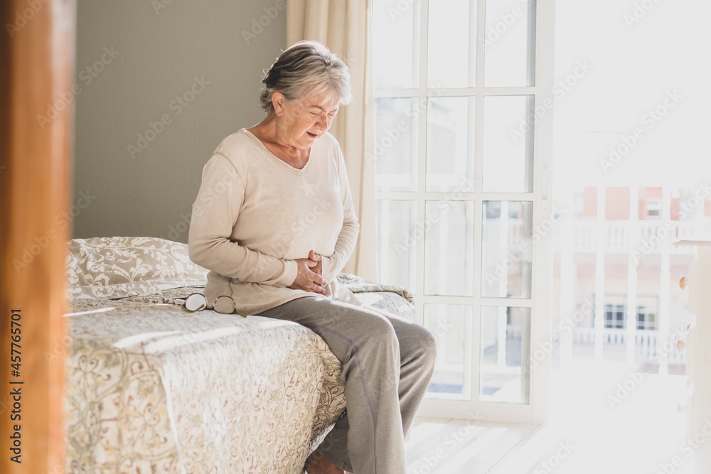 Senior woman holding belly suffering from abdomen pain in morning on bed at home. Old retired female sitting on bed touching her stomach feeling discomfort in bedroom at home Senior woman holding belly suffering from abdomen pain in morning on bed at home. Old retired female sitting on bed touching her stomach feeling discomfort in bedroom at home