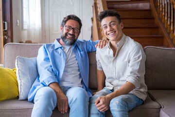 Happy father and son sitting on sofa at home. Cheerful father with teenage son sitting on couch in the living room of apartment. Portrait of father and son spending leisure time at home