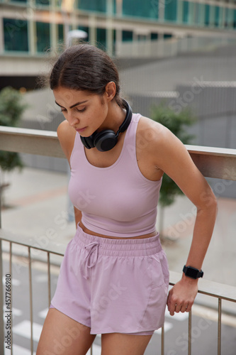 Motivation and healthy lifestyle concept. Fit slim young woman rests after cardio workout focused down dressed in sportswear uses headphonnes and smartwatch trains outdoors works on slimming