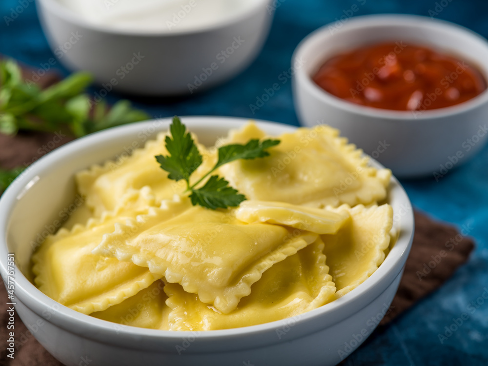 Serving Italian ravioli in a deep white bowl, topped with a green mint leaf. In the background there are two sauces and sour cream. Restaurant, hotel. cafe, home cooking, menu design. Serving Italian ravioli in a deep white bowl, topped with a green mint leaf. In the background there are two sauces and sour cream. Restaurant, hotel. cafe, home cooking, menu design.
