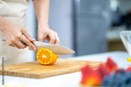 Asian woman bakery shop owner preparing bakery in the kitchen. Adult female using kitchen knife cut fresh fruit for making fruit tart. Small business entrepreneur and indoor lifestyle baking concept.