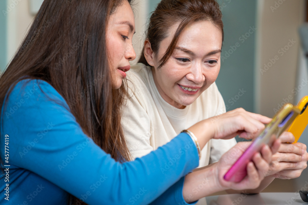 Millennial Asian woman friends sitting on sofa in living room and playing games on smartphone together. Modern female friendship enjoy weekend activity lifestyle with wireless technology at home.