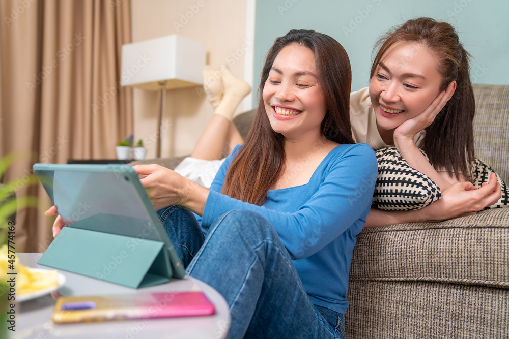 Asian woman friends sitting in living room using digital tablet for watching movie or social media together. Modern female friendship enjoy weekend activity lifestyle with wireless technology at home. Asian woman friends sitting in living room using digital tablet for watching movie or social media together. Modern female friendship enjoy weekend activity lifestyle with wireless technology at home.