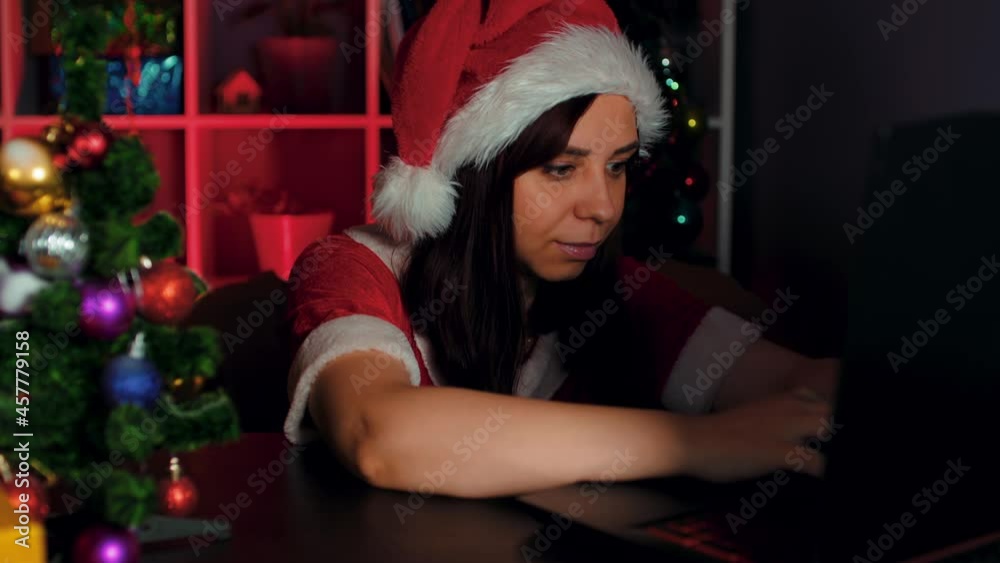 Young woman in santa costume works at computer in office. Employee sitting in armchair and working at laptop in New Year's atmosphere. Young woman in santa costume works at computer in office. Employee sitting in armchair and working at laptop in New Year's atmosphere.