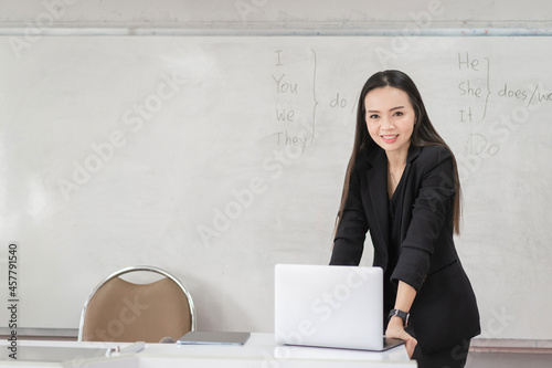 Adult confident cheerful Asian woman teacher in business uniform with digital tablet and laptop stands in front of whiteboard teaching modern in classroom