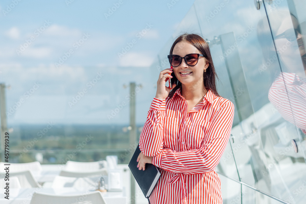 A young dark-haired businesswoman with a notebook in her hands is talking on a smartphone.Business negotiations on the outdoor terrace of the cafe on a summer day.Business concept.Copy space for text. A young dark-haired businesswoman with a notebook in her hands is talking on a smartphone.Business negotiations on the outdoor terrace of the cafe on a summer day.Business concept.Copy space for text.