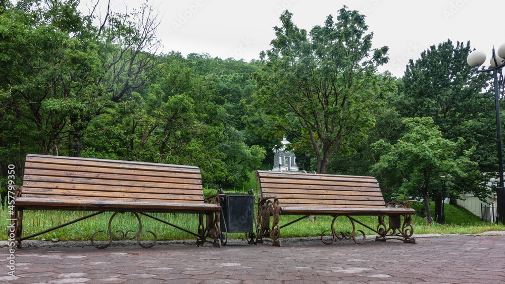 There are wooden benches on the paved path in the park. Behind - a lawn, green trees. An old white chapel is visible among the foliage. Petropavlovsk-Kamchatsky