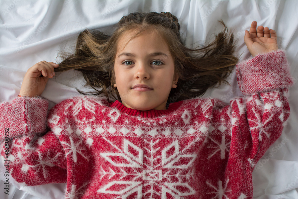 Little Girl With Positive And Joyful Attitude At Christmas. 6-Year-Old Girl Dressed In Christmas Clothes In The Bedroom At Home Lying On The Bed And Looking At The Camera. White Background Little Girl With Positive And Joyful Attitude At Christmas. 6-Year-Old Girl Dressed In Christmas Clothes In The Bedroom At Home Lying On The Bed And Looking At The Camera. White Background