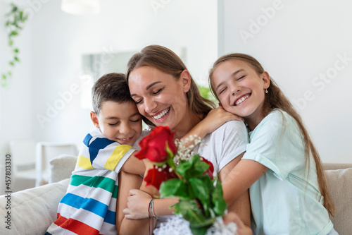 Young mother with a bouquet of roses laughs, hugging her son, and сheerful girl with a card congratulates mom during holiday celebration in kitchen at home