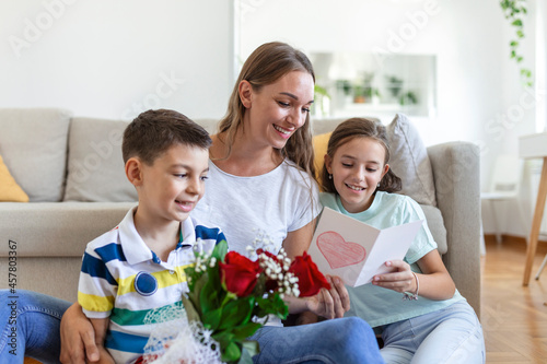 Young mother with a bouquet of roses laughs, hugging her son, and сheerful girl with a card and roses congratulates mom during holiday celebration in kitchen at home