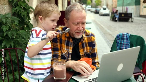 Cheerful happy little boy with grandfather using laptop computer outside in cafe enjoying beautiful sunny day. Freelance urban lifestyle. Joyful adult bearded man talking with grandson. Internet 