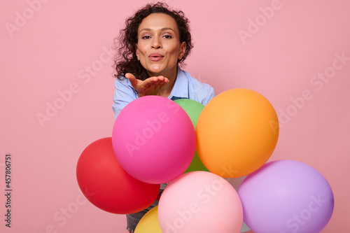 Attractive African American woman sending an air kiss to the camera, standing behind multi-colored balloons on pink background with copy space. Beautiful mixed race lady with colorful air balls