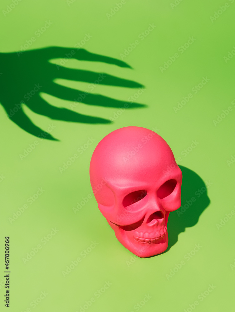 Pink human skull with a spooky hand shadow catching it from behind on a neon green background. Creative Halloween party concept. Pop art invitation card visual trend. Pink human skull with a spooky hand shadow catching it from behind on a neon green background. Creative Halloween party concept. Pop art invitation card visual trend.