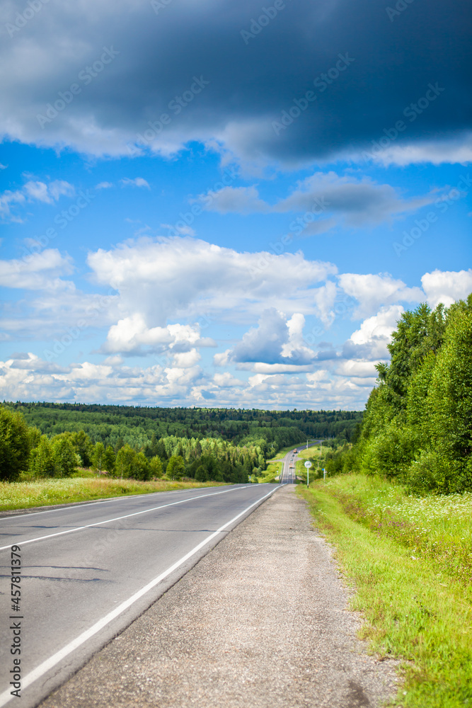 Straight road with a marking on the nature background. Open Road in future, no cars, auto on asphalt road through green forest, trees. Clouds on blue sky in summer, sunshine, sunny day. Bottom view