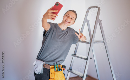 Beautiful young happy woman repairman standing by a stepladder in a belt with tools taking a selfie photo on a smartphone © olezzo