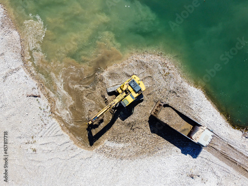 Excavator digging and loading sand into dump truck at river bank. Heavy machinery working at sand quarry. Aerial drone view of digger and truck loading gravel in nature.Environment and water pollution