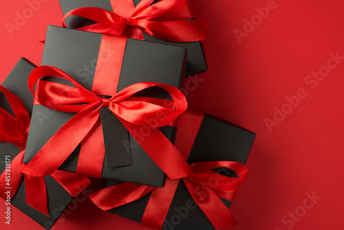 Top view photo of stack of black gift boxes with red ribbon bow and tag on isolated red background with empty space