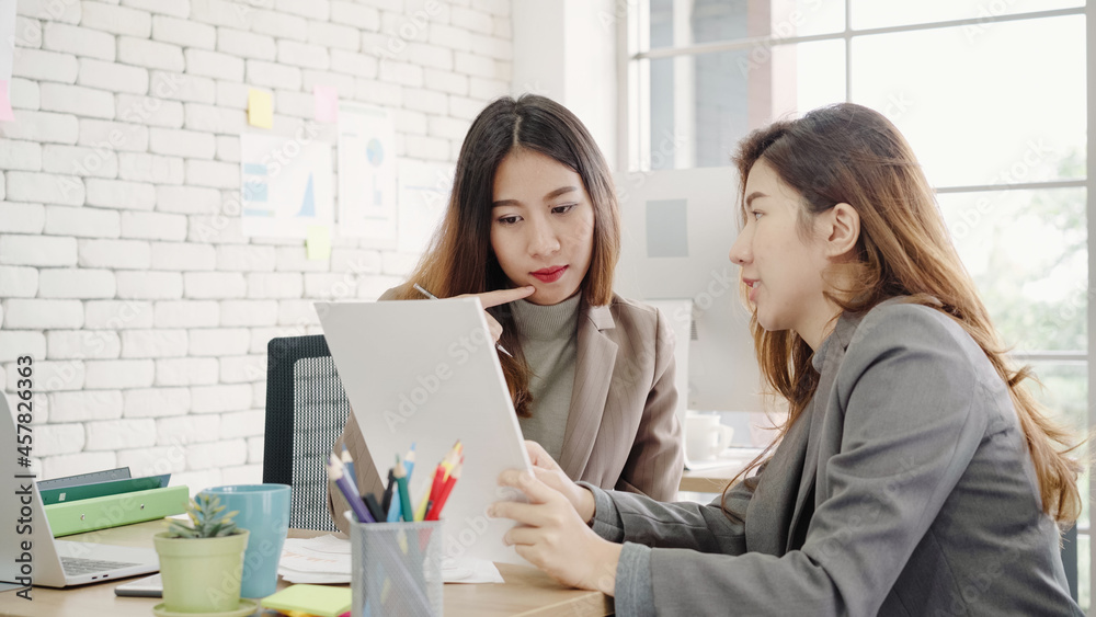 Two young Asian businesswomen working together in office at small business sitting reading a report or paperwork with pleased smiles and successful teamwork. Business, technology and office concept. Two young Asian businesswomen working together in office at small business sitting reading a report or paperwork with pleased smiles and successful teamwork. Business, technology and office concept.