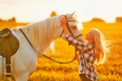 Young cute happy joyful satisfied smiling woman hugging and stroking beautiful white horse at meadow at sunset at golden hour