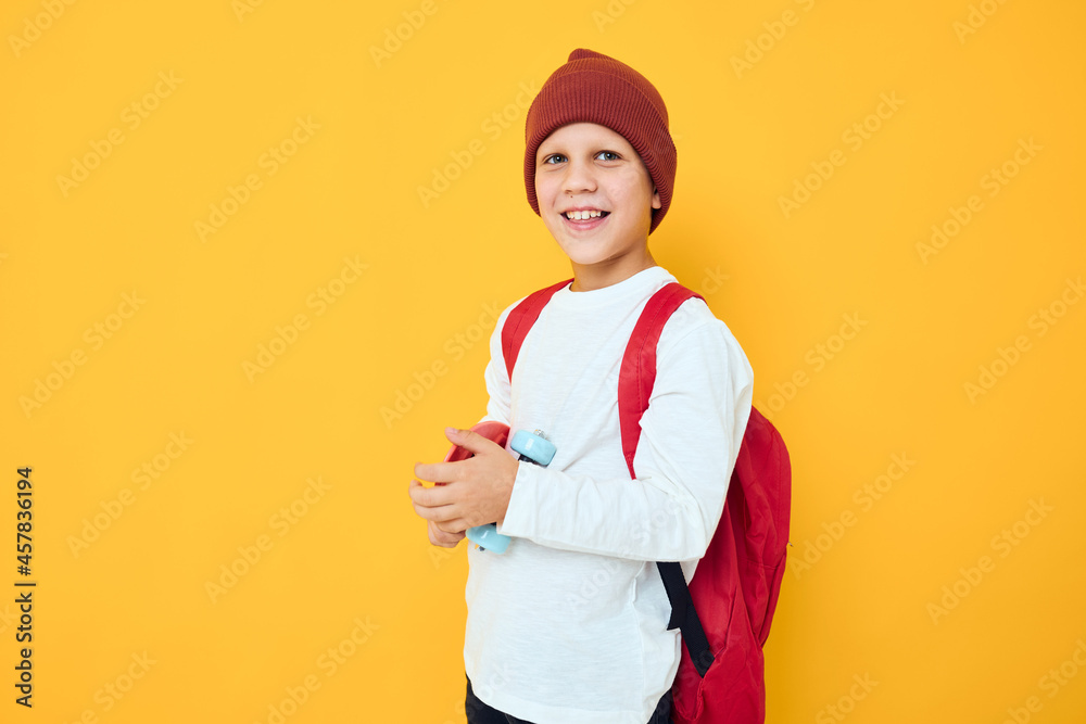 Cheerful little kid with red backpack red skateboard yellow color background Cheerful little kid with red backpack red skateboard yellow color background