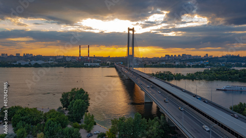 South bridge in Kiev. Sunset over the Dnieper. Thick clouds over the evening city. Evening shot of the bridge. Orange sun at sunset. The rays break through the clouds and are reflected in the river.