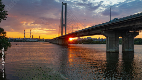 South bridge in Kiev. Sunset over the Dnieper. Thick clouds over the evening city. Evening shot of the bridge. Orange sun at sunset. The rays break through the clouds and are reflected in the river.