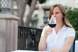 A young stylish beautiful blonde woman in a white shirt is drinking red wine on the open summer veranda of the Vodino restaurant in the evening. A beautiful look and a light playful smile