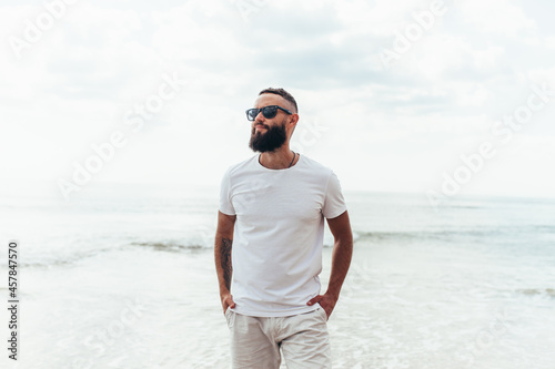 Young bearded hipster guy with beard in white t-shirt and sunglasses at the tropical beach. Mock-up for print. T-shirt template.