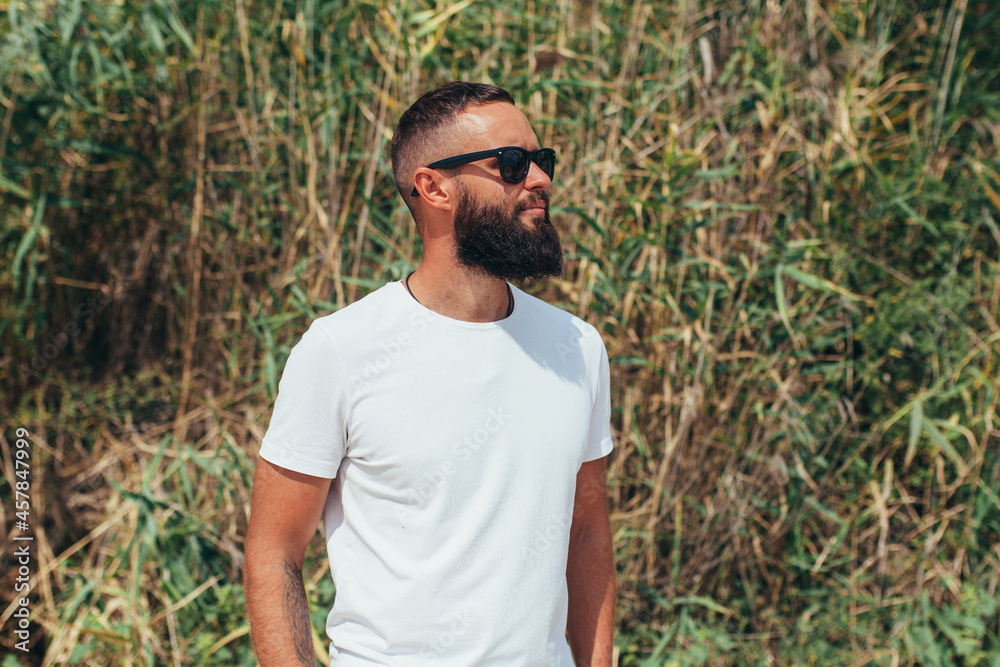 Young bearded hipster guy with beard in white t-shirt and sunglasses at the tropical beach. Mock-up for print. T-shirt template. Young bearded hipster guy with beard in white t-shirt and sunglasses at the tropical beach. Mock-up for print. T-shirt template.