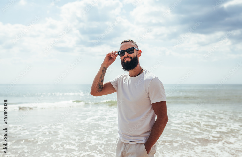 Young bearded hipster guy with beard in white t-shirt and sunglasses at the tropical beach. Mock-up for print. T-shirt template. Young bearded hipster guy with beard in white t-shirt and sunglasses at the tropical beach. Mock-up for print. T-shirt template.