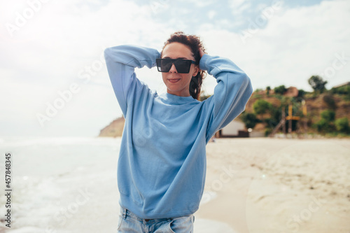 Beautiful young adult woman in blue blank sweatshirt and sunglasses on a background of a tropical beach. Mock-up for print. Space for your logo or design.