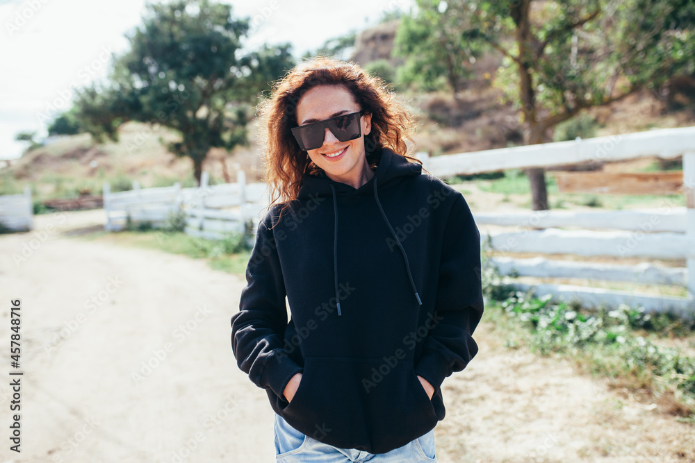 Beautiful young adult woman in black hoodie or sweatshirt and sunglasses against the background of a wooden fence and nature. Mock-up for print. Space for your logo or design. Beautiful young adult woman in black hoodie or sweatshirt and sunglasses against the background of a wooden fence and nature. Mock-up for print. Space for your logo or design.