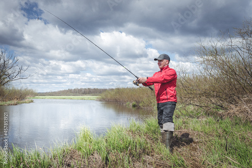 Fisherman in a red jacket is fishing on the green bank of the river.