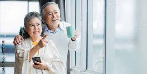portrait of Asian senior retired couple smiling and looking out of window apartment while hold wife shoulder .Cheerful asian senior couple retirement life. wellness Senior healthy lifestyle concept.