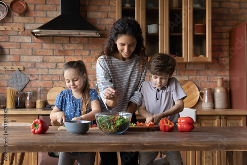 Happy affectionate millennial mother involved in preparing healthy vegetarian food with adorable small preschool children siblings  teaching cooking in old-fashioned kitchen  culinary concept.
