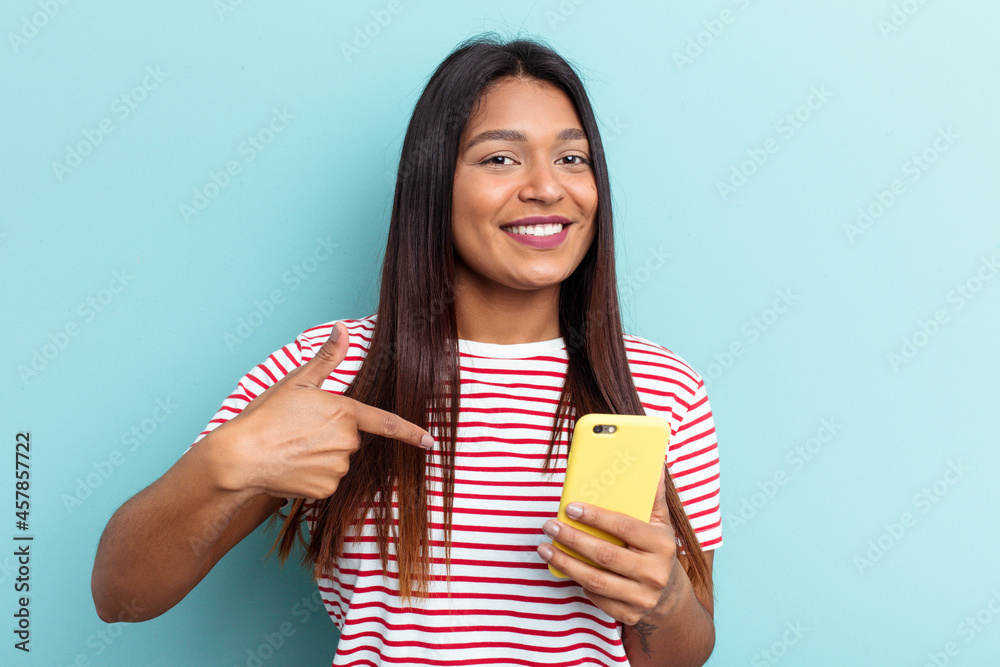 Young Venezuelan woman holding mobile phone isolated on blue background person pointing by hand to a shirt copy space, proud and confident Young Venezuelan woman holding mobile phone isolated on blue background person pointing by hand to a shirt copy space, proud and confident